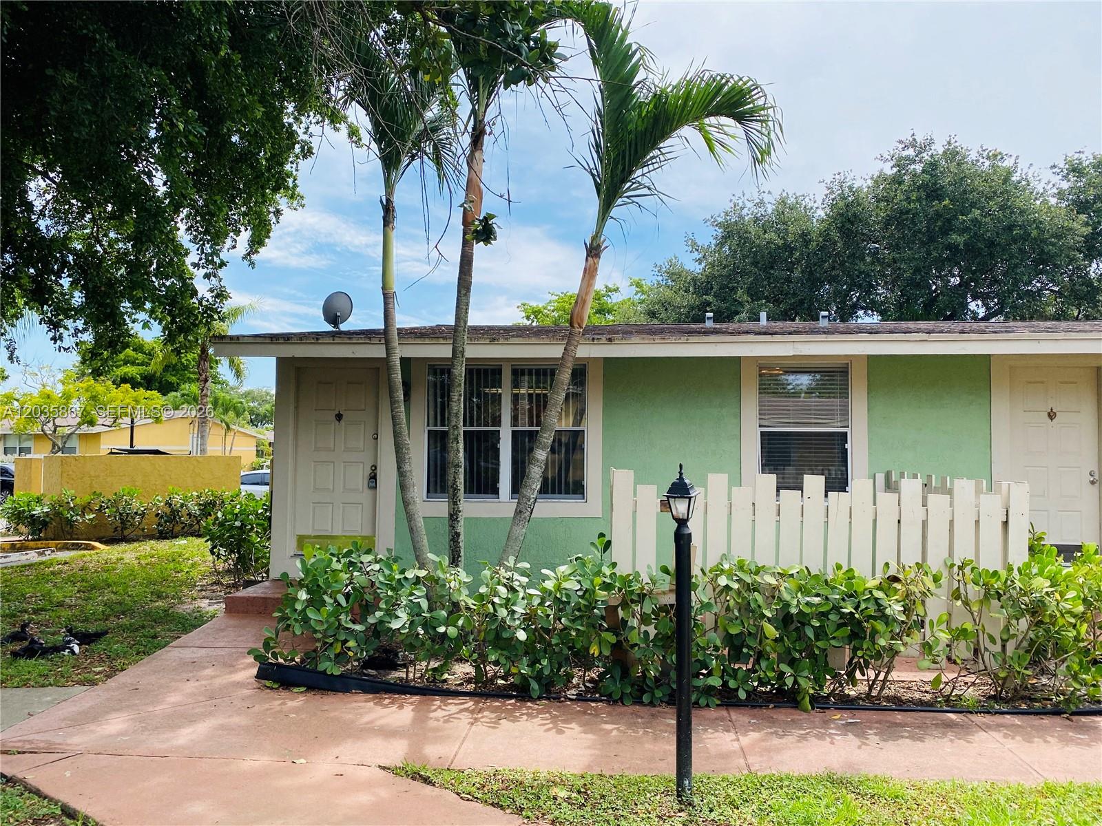 The exterior of a light green house features a white fence, palm trees, and walkways — TAMARAC, FL