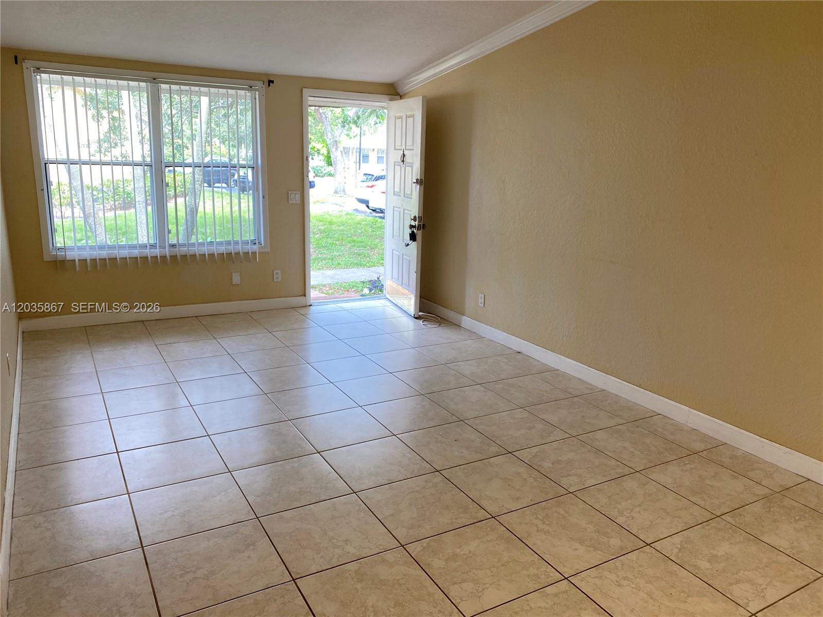 An empty room features beige walls, light-colored tile flooring, a large window, and an open door — TAMARAC, FL