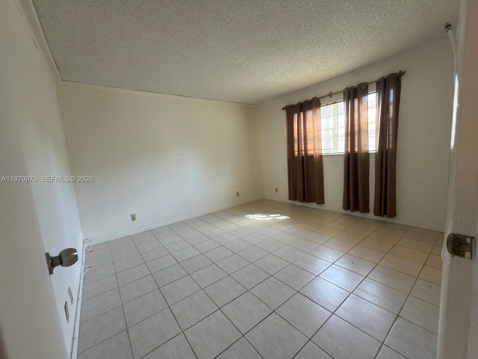An empty room features white walls, light-colored tile flooring, and brown curtains over a window — 30 SE 4th Ave # 103, Hallandale Beach FL 33009, HALLANDALE BEACH, FL