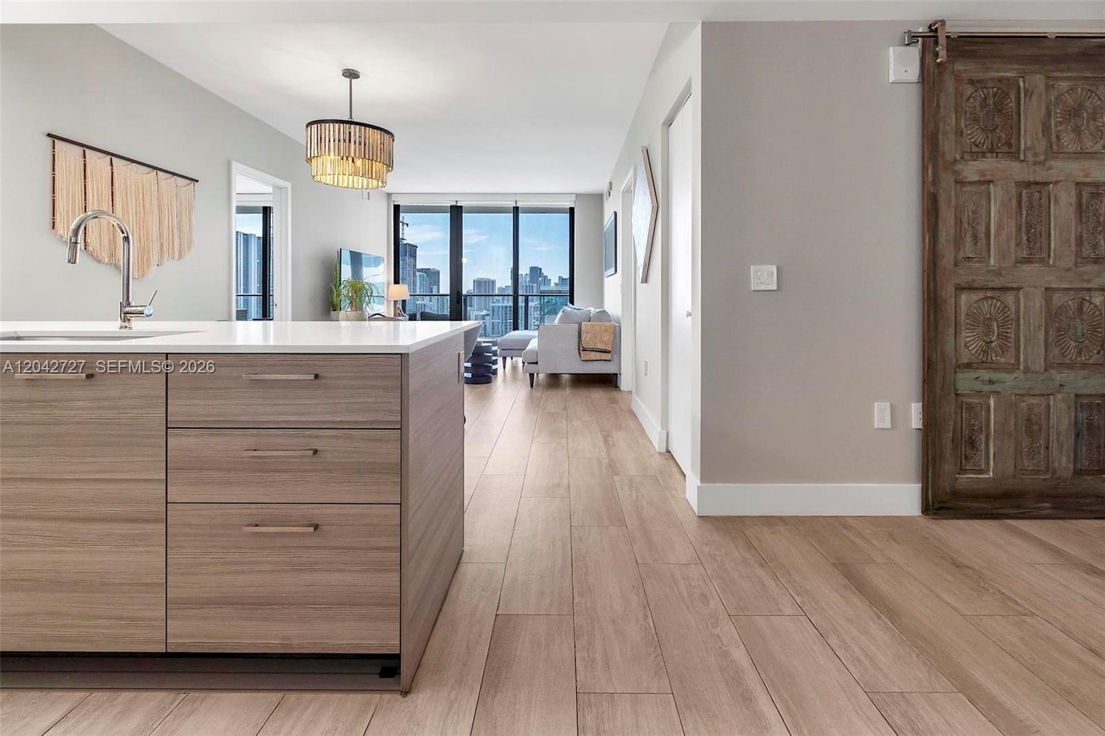 A modern kitchen island with wood-grain cabinets sits before a living area with city views — 121 NE 34th St # 2502, Miami FL 33137, MIAMI, FL