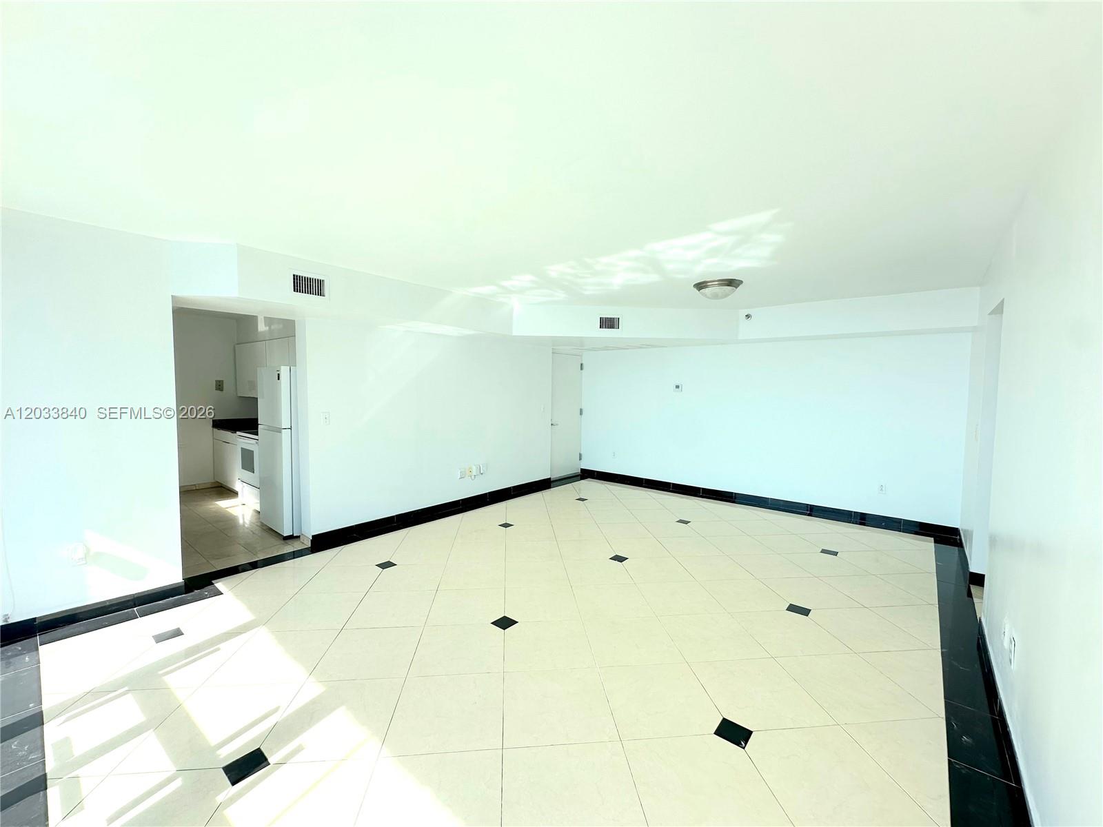 An empty room features white walls, light-colored diagonal floor tiles with black accents, and an adjacent kitchen — 5900 Collins Ave # 906, Miami Beach FL 33140, MIAMI BEACH, FL