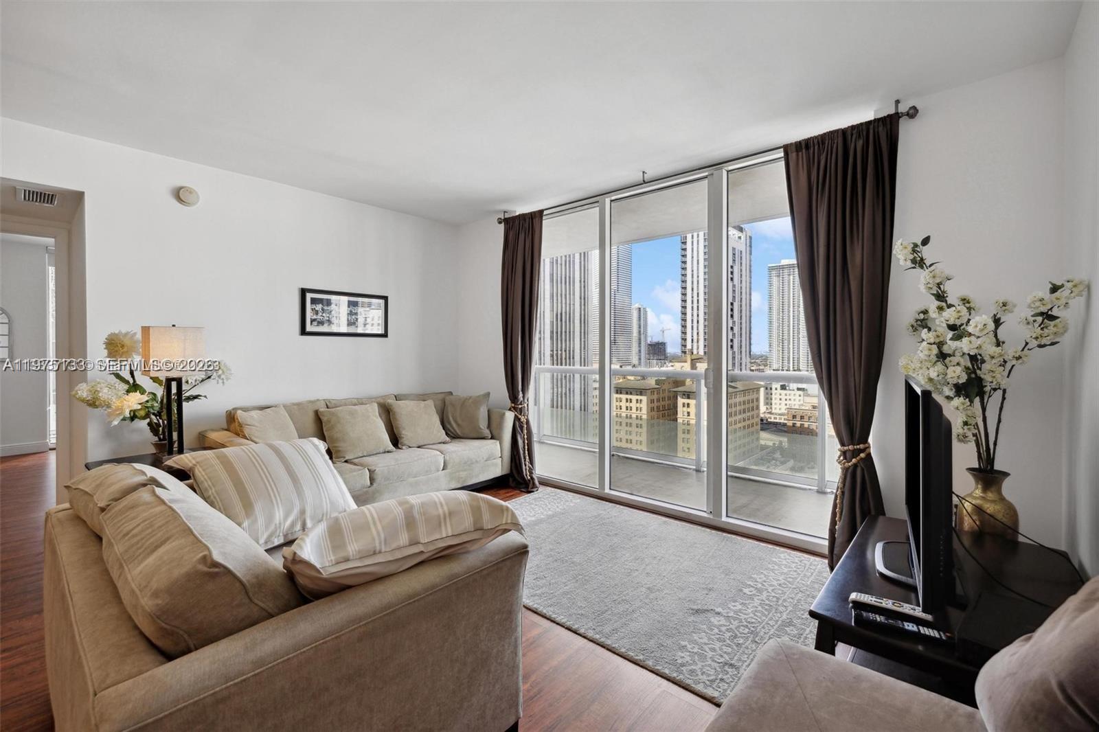 A living room features a beige sofa, a television, and floor-to-ceiling windows with city views — 50 Biscayne Blvd # 2111, Miami FL 33132, MIAMI, FL