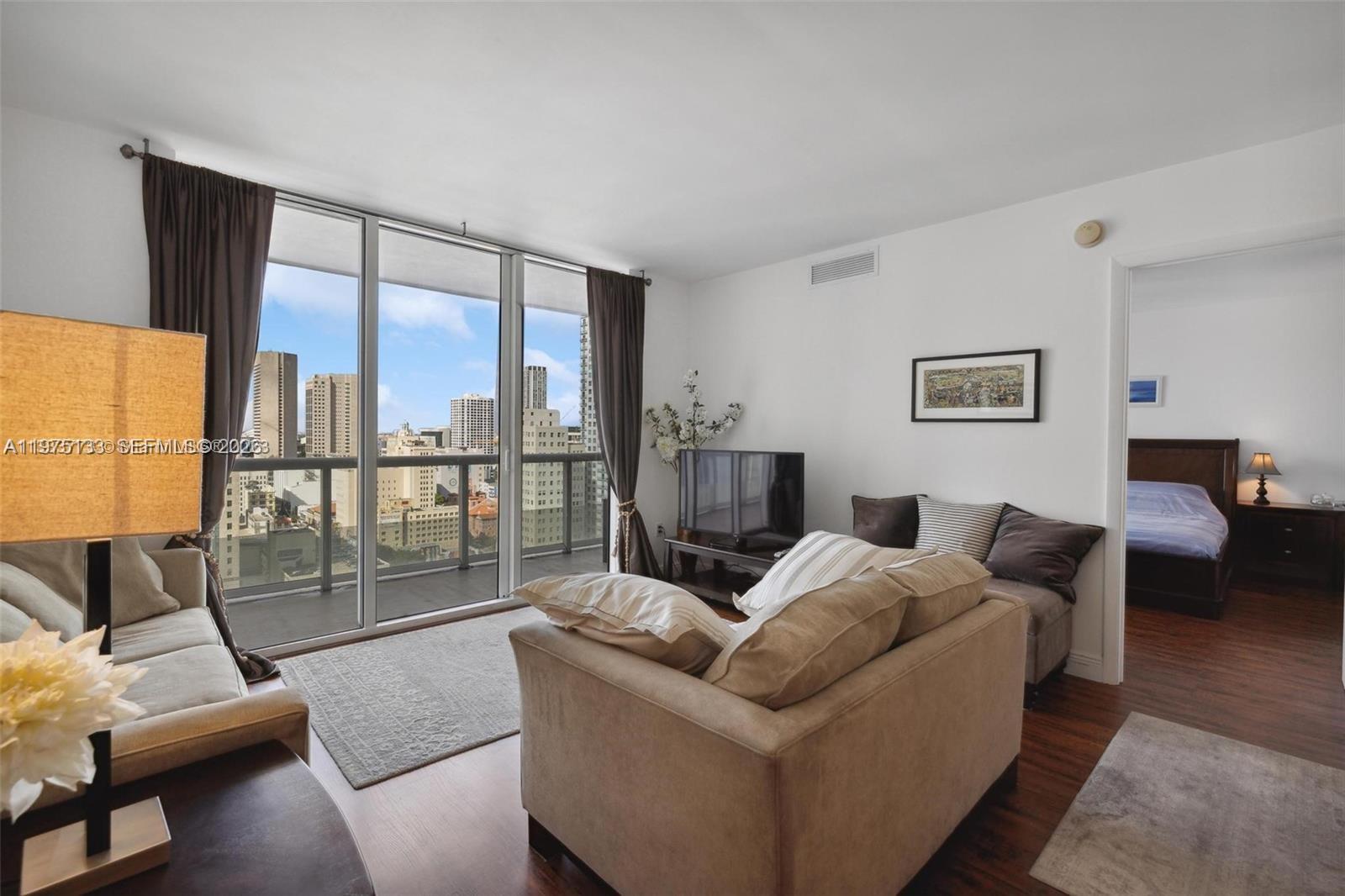 A living room with a beige sofa, floor-to-ceiling windows, and a view of city buildings — 50 Biscayne Blvd # 2111, Miami FL 33132, MIAMI, FL