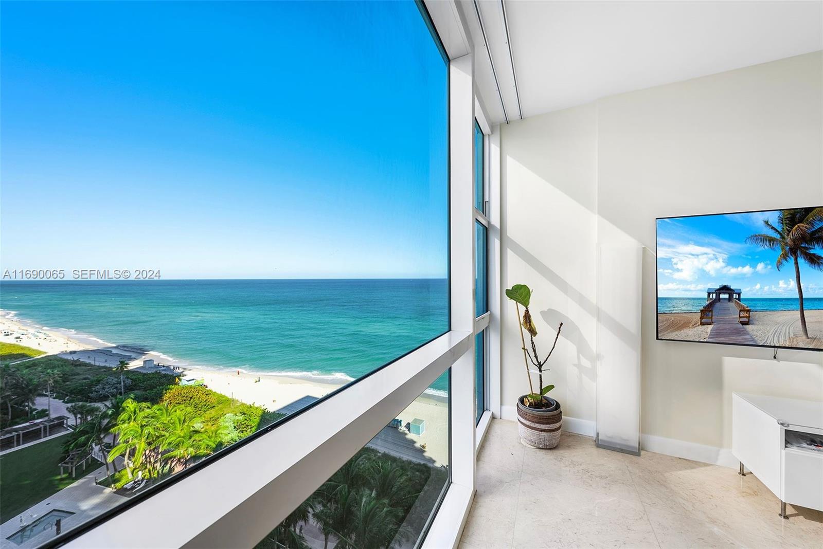 A room with floor-to-ceiling windows overlooks a beach and ocean, featuring a wall-mounted television — 6801 Collins Ave # 1203, Miami Beach FL 33141, MIAMI BEACH, FL