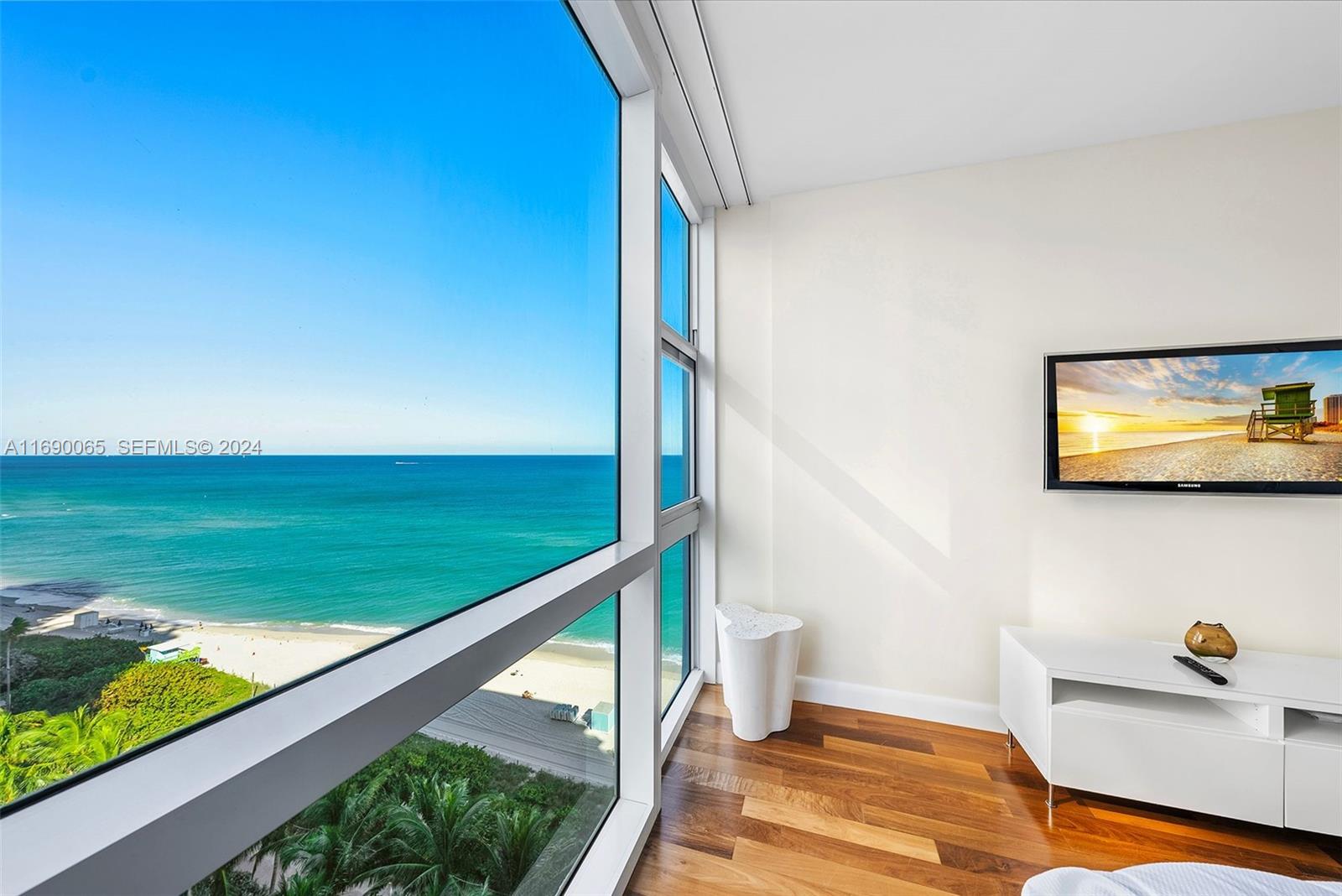 A room with hardwood floors features a wall-mounted television and floor-to-ceiling windows overlooking the ocean — 6801 Collins Ave # 1203, Miami Beach FL 33141, MIAMI BEACH, FL