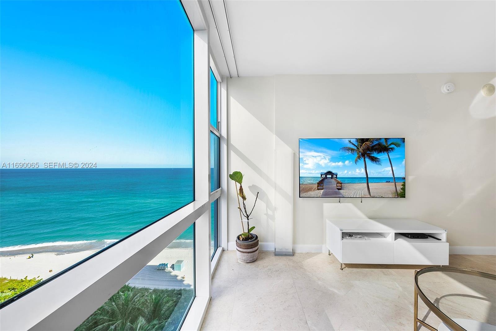 A room with a large window overlooking the ocean, a wall-mounted television, and tile flooring — 6801 Collins Ave # 1203, Miami Beach FL 33141, MIAMI BEACH, FL