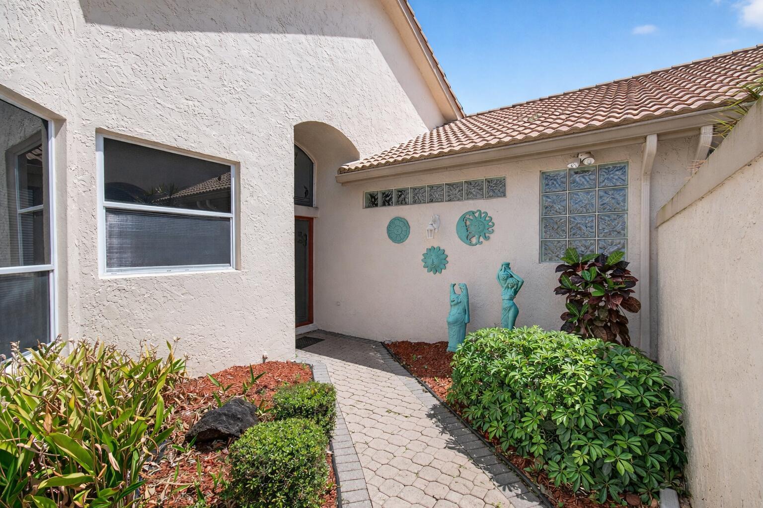 A paved walkway leads to the front entrance of a stucco home with decorative wall art — 8864 Shoal Creek Lane, Boynton Beach FL 33472, BOYNTON BEACH, FL