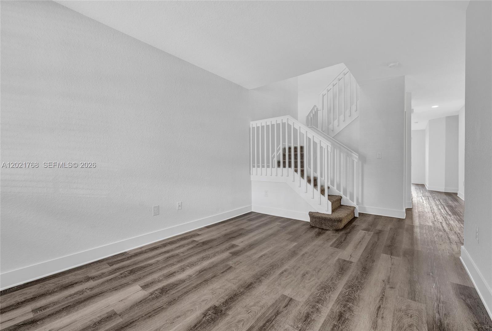 An empty room features wood-look flooring, white walls, and a staircase with white railings — 1445 SE 24th Rd, Homestead FL 33035, HOMESTEAD, FL