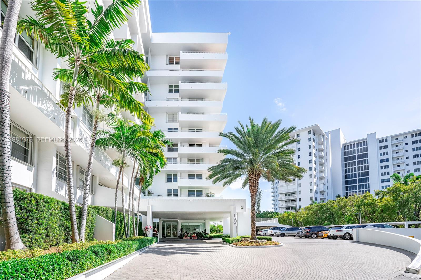 A white multi-story apartment building features a paved driveway, palm trees, and a parking area — 199 Ocean Lane Dr # 1207, Key Biscayne FL 33149, KEY BISCAYNE, FL