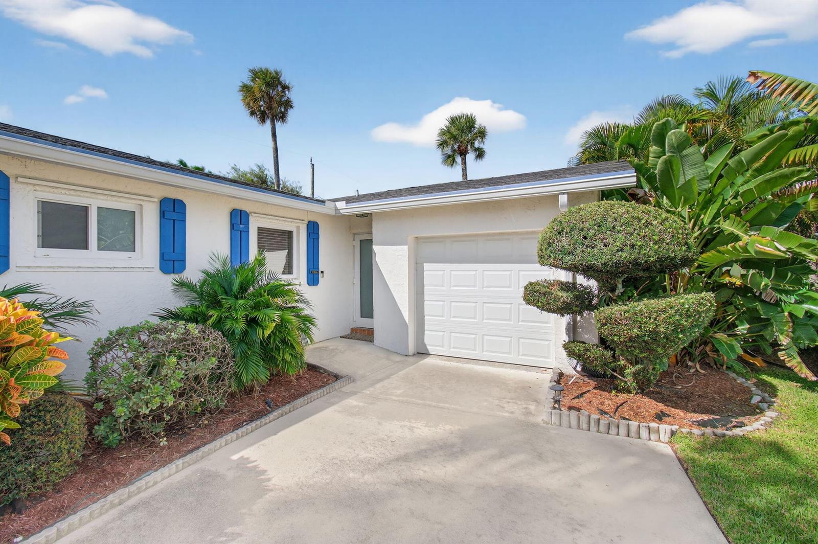 A white house exterior features a single-car garage, blue shutters, and tropical landscaping — 802 Mallard Drive, Delray Beach FL 33444, DELRAY BEACH, FL