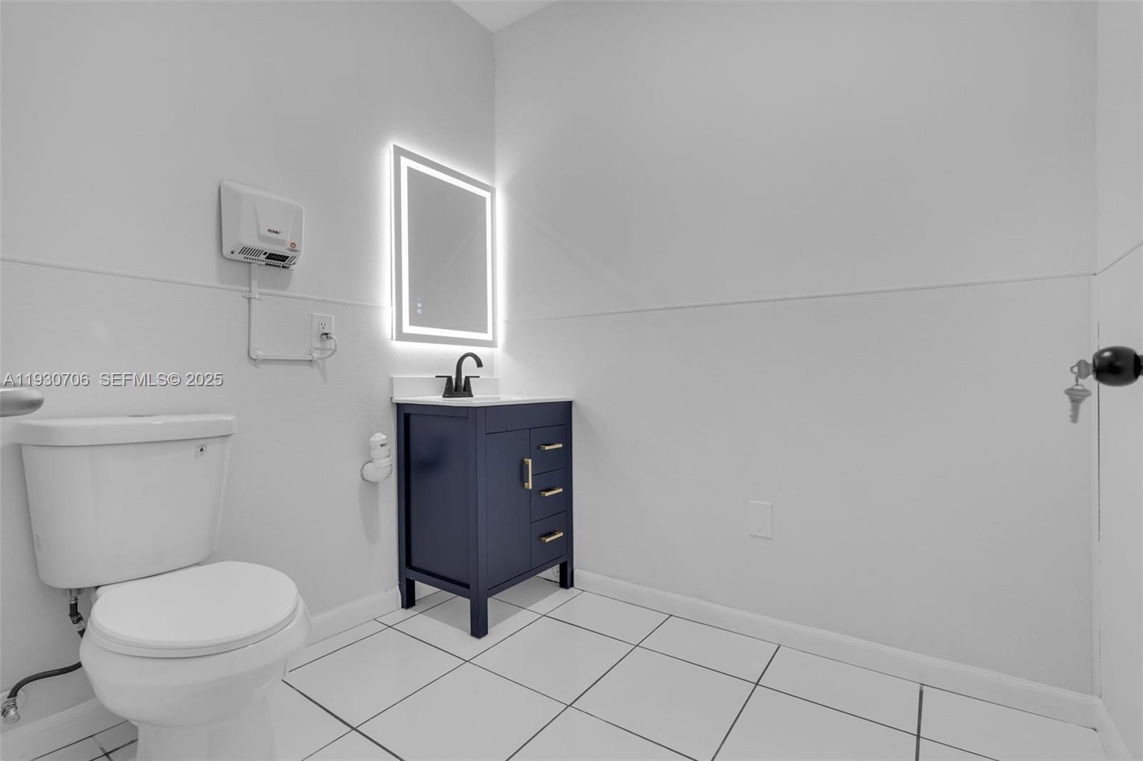 A small bathroom features a white toilet, a dark blue vanity, and an illuminated mirror — 8930 SW 40th St, Miami FL 33165, MIAMI, FL