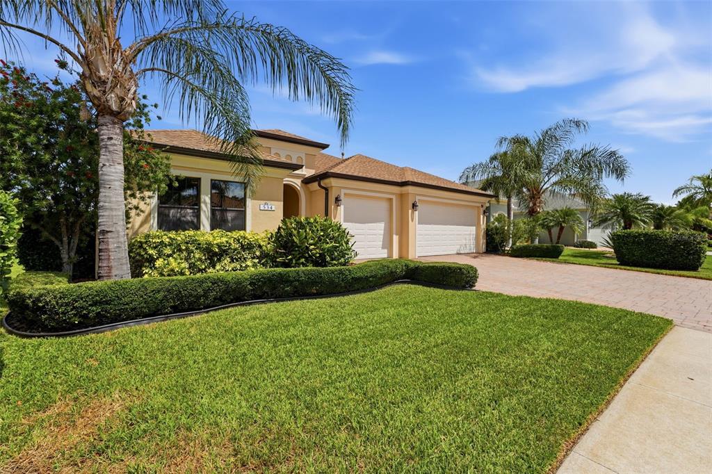A single-story house with a tan exterior, two-car garage, and manicured landscaping under a blue sky — 534 CHANTILLY TRAIL, BRADENTON, FL