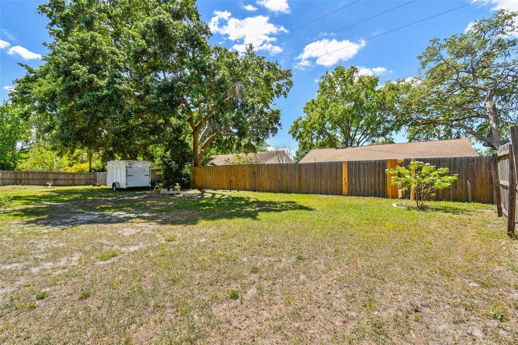 A grassy backyard features a wooden fence, a white utility trailer, and several large trees — 24231 SATINWOOD COURT, LUTZ, FL