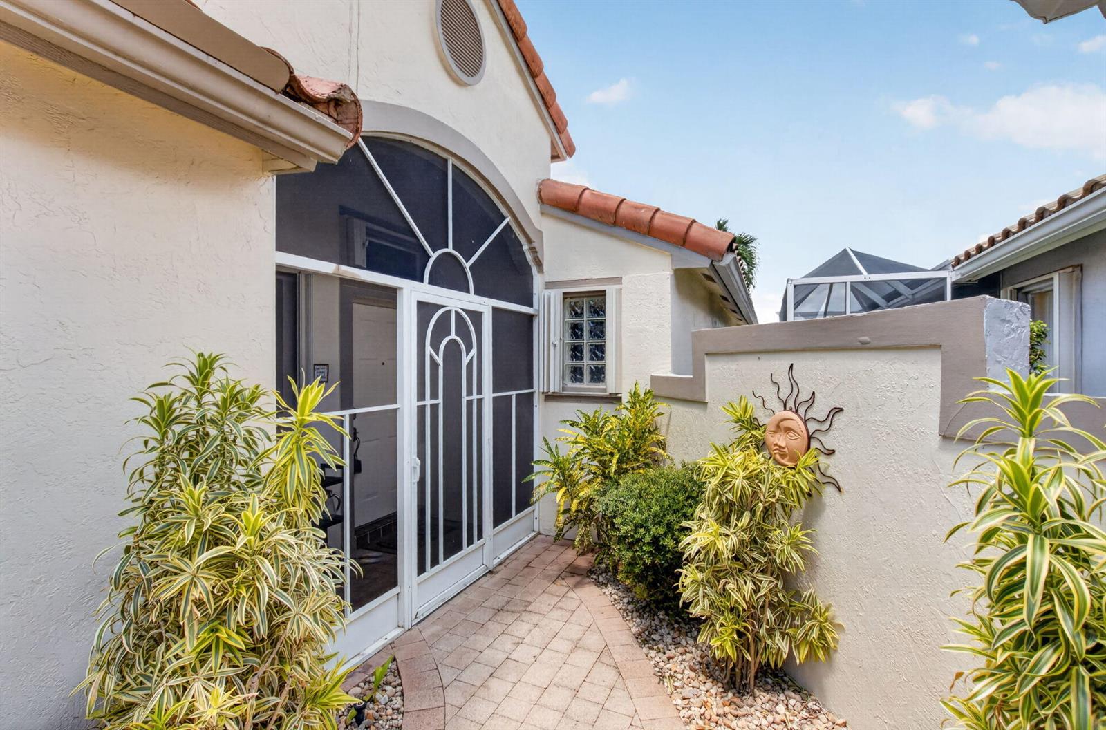 A paved walkway leads to a screened entryway beside a stucco wall with sun decor — 11585 Augustus Drive, Boynton Beach FL 33437, BOYNTON BEACH, FL