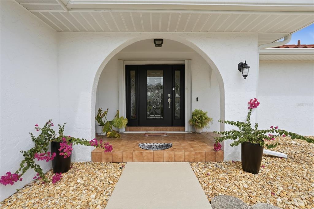 A white arched entryway features a black front door, tiled porch, and potted bougainvillea plants — 3433 NIGHTHAWK COURT, PUNTA GORDA, FL