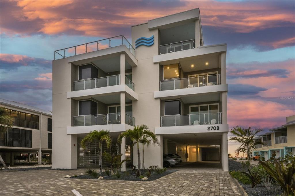 A modern three-story building with balconies and covered parking stands under a sunset sky — 2708 N BEACH ROAD UNIT 3, ENGLEWOOD, FL