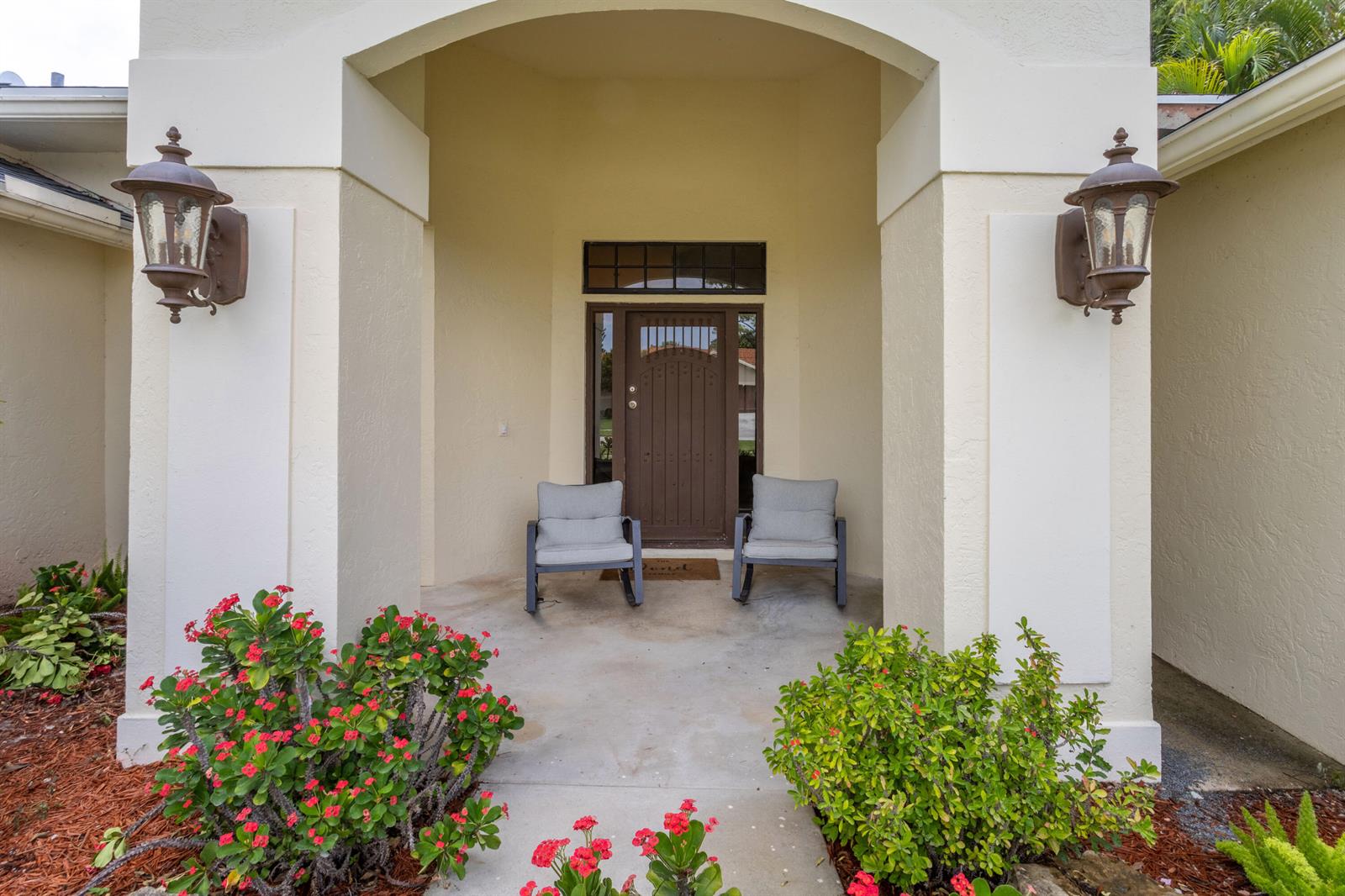 A covered entryway features a brown front door, two gray chairs, and two wall-mounted lanterns — 13823 Callington Drive, Wellington FL 33414, WELLINGTON, FL