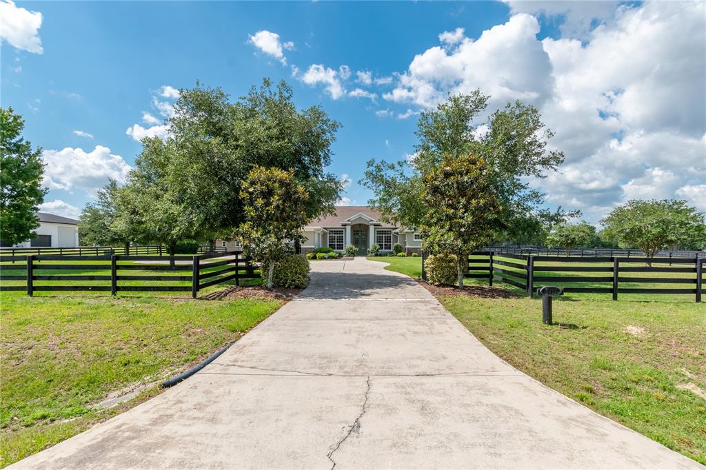 A concrete driveway leads to a single-story house framed by trees and black wooden fencing — 6957 NW 54TH LOOP, OCALA, FL