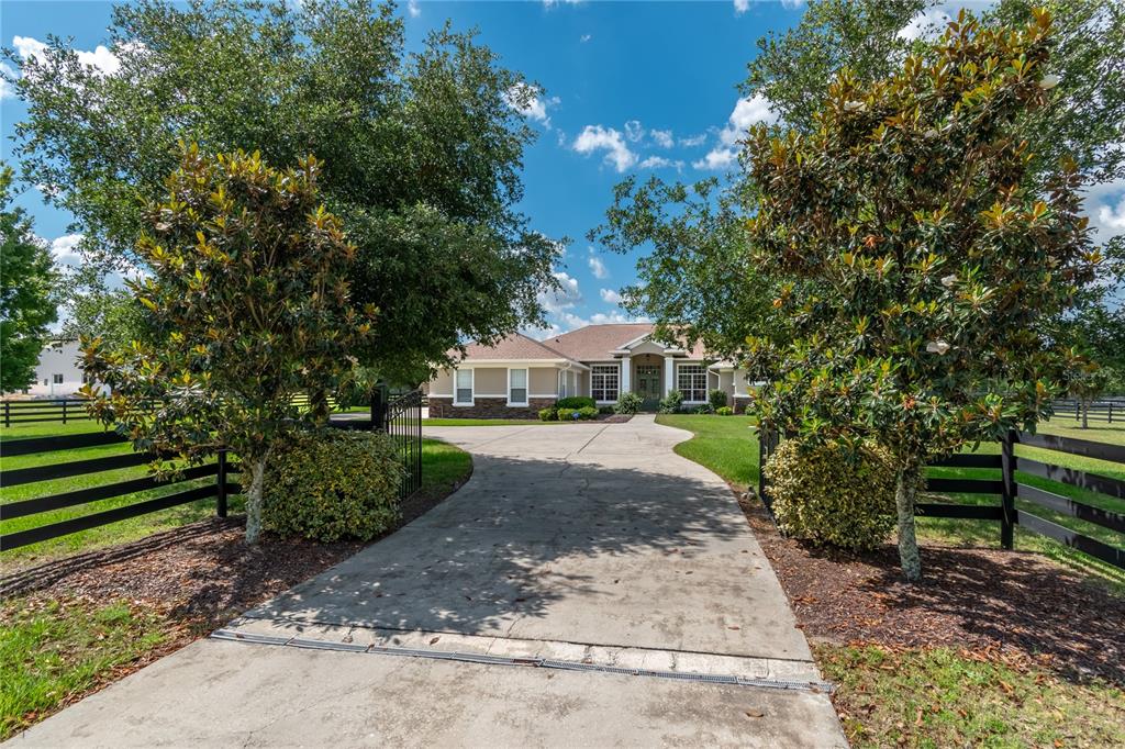 A concrete driveway leads to a single-story house framed by trees and black wooden fencing — 6957 NW 54TH LOOP, OCALA, FL