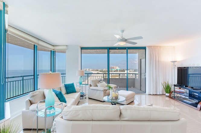 A bright living room features white leather sofas, a glass-topped table, and floor-to-ceiling windows overlooking the ocean — 1520 GULF BOULEVARD UNIT 1602, CLEARWATER BEACH, FL