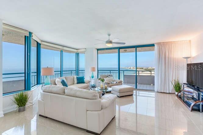 A bright living room features white sofas, tile floors, and floor-to-ceiling windows overlooking the ocean — 1520 GULF BOULEVARD UNIT 1602, CLEARWATER BEACH, FL