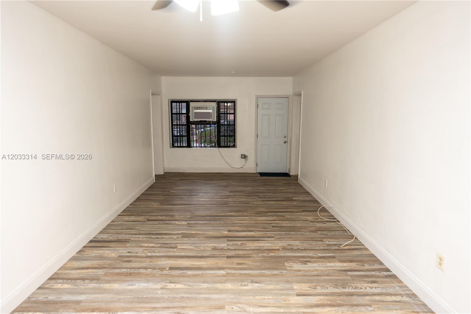 An empty room features white walls, wood-look flooring, a window with a security grate, and a door — 259 NE 56th St, Miami FL 33137, MIAMI, FL