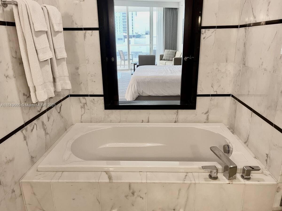 A white soaking tub is set against marble-tiled walls with a large mirror above — 4391 Collins Ave # 918, Miami Beach FL 33140, MIAMI BEACH, FL