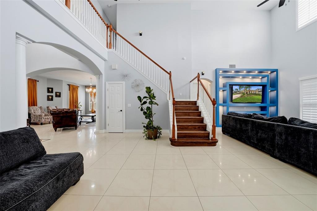 A spacious living room features a grand staircase, tile flooring, and a blue entertainment unit — 18043 COZUMEL ISLE DRIVE, TAMPA, FL