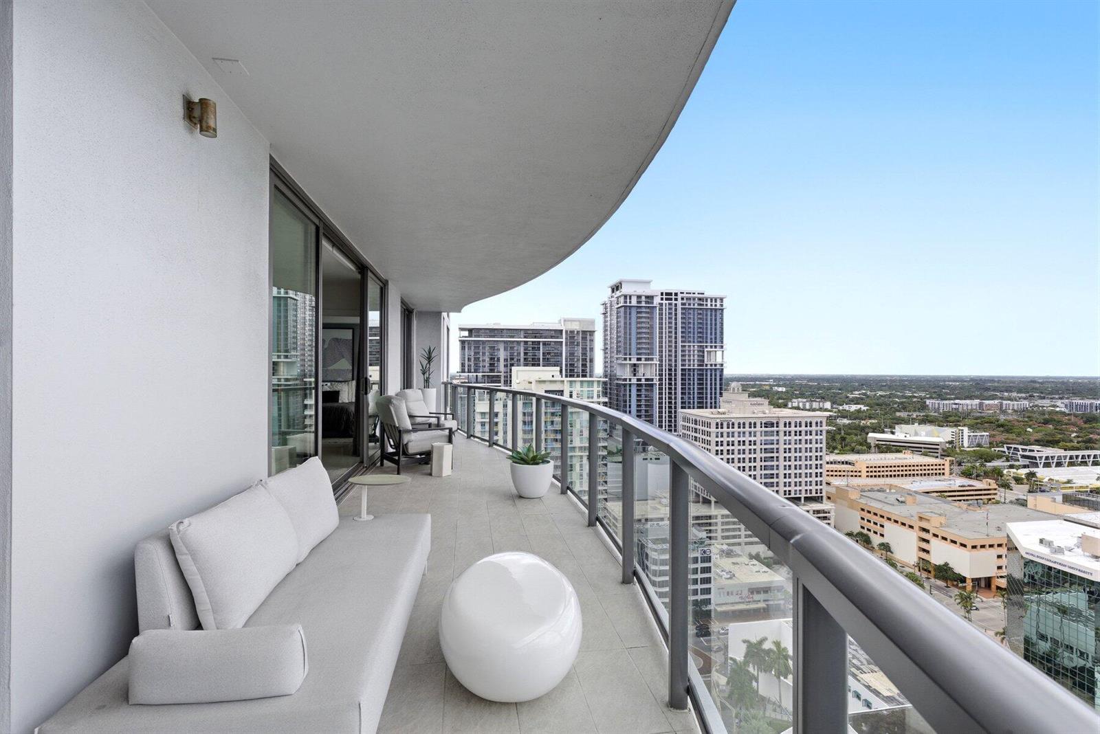 A curved balcony with light-colored furniture overlooks a city skyline with tall buildings — 100 E Las Olas Boulevard # 2802, Fort Lauderdale FL 33301, FORT LAUDERDALE, FL