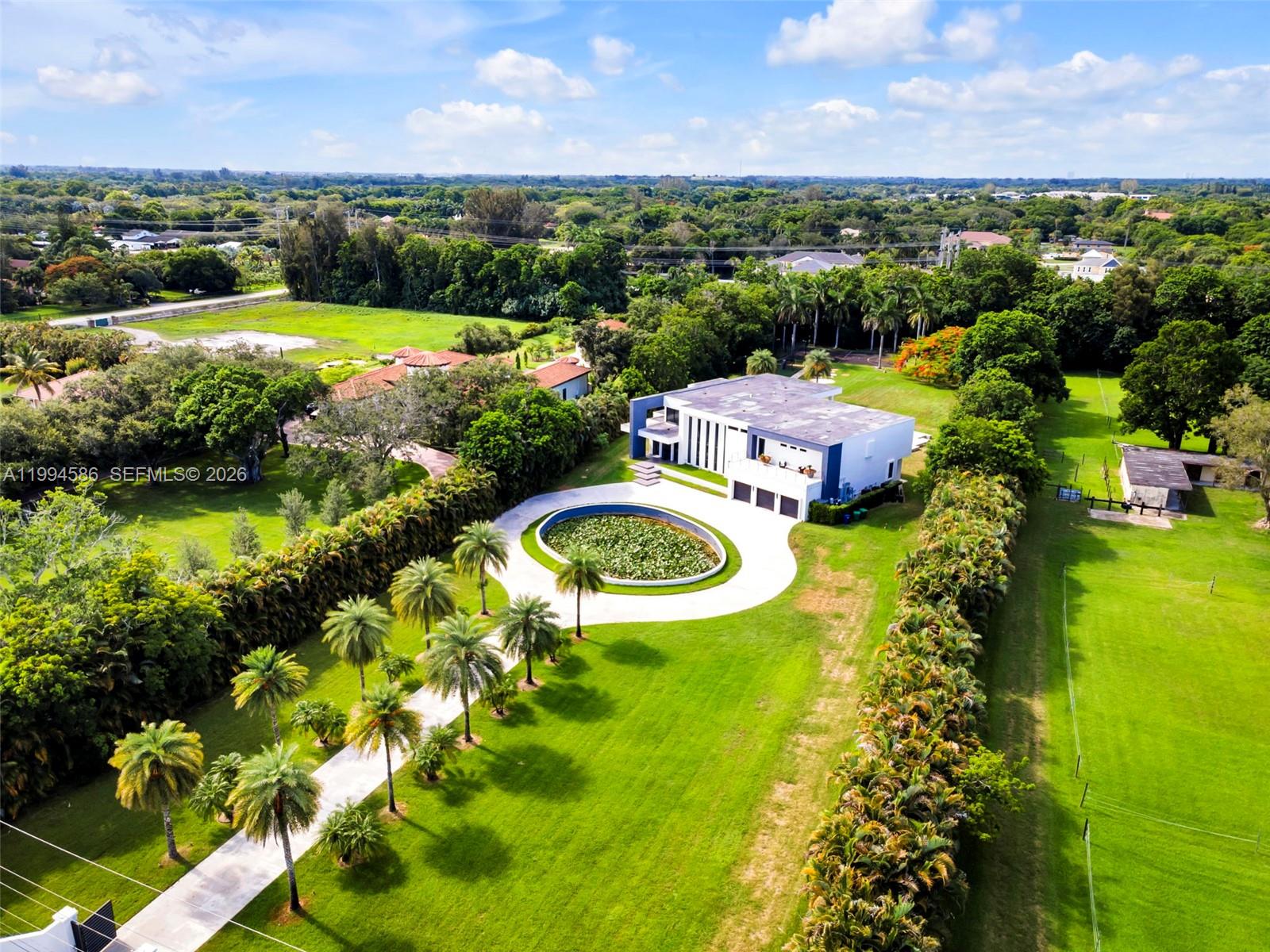 An aerial view shows a modern house with a circular pond and palm-lined driveway — 13441 Mustang Trl, Southwest Ranches FL 33330, SOUTHWEST RANCHES, FL