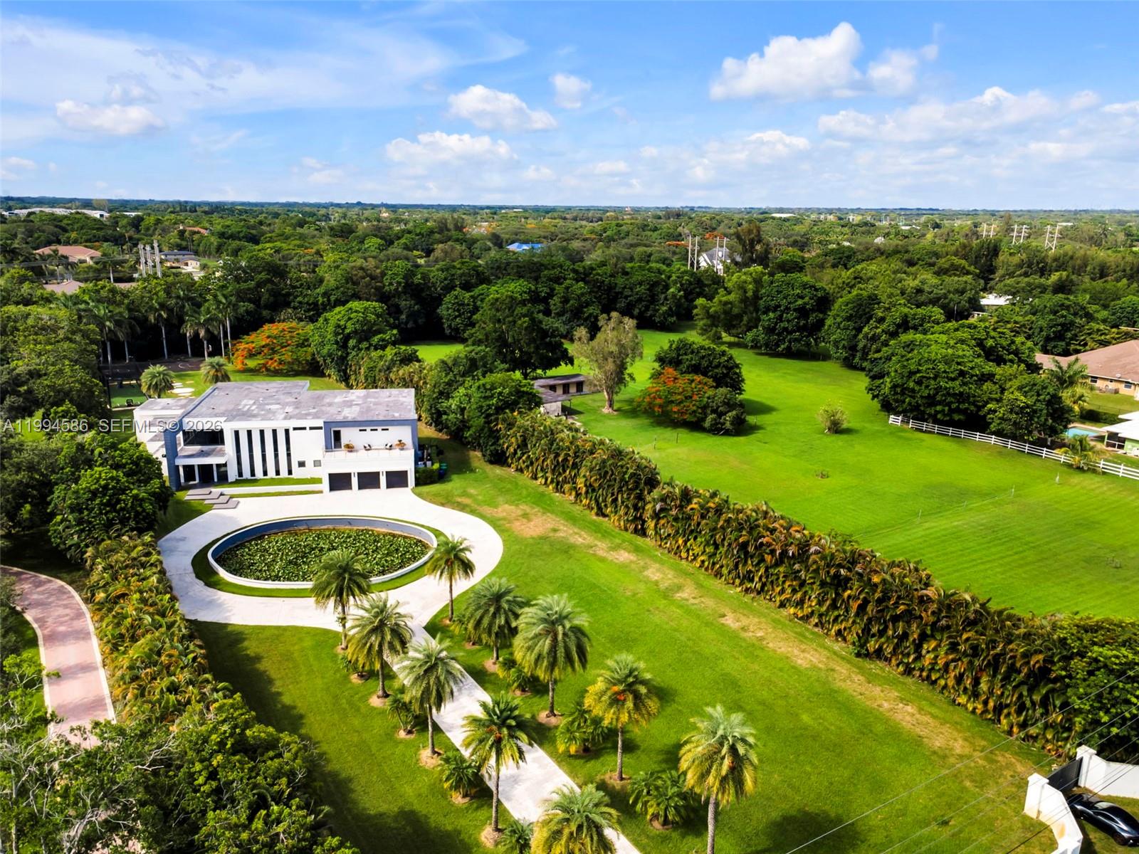 An aerial view shows a modern house with a circular pond and palm-lined driveway — 13441 Mustang Trl, Southwest Ranches FL 33330, SOUTHWEST RANCHES, FL