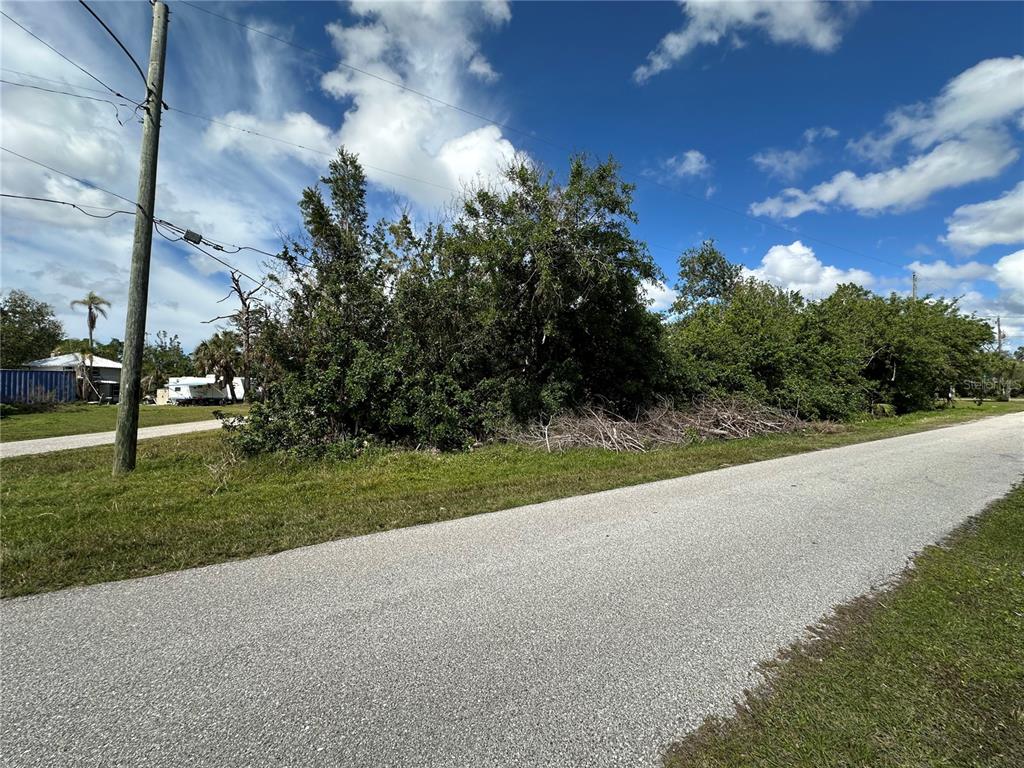 A paved road runs alongside a vacant, tree-covered lot under a blue sky — 4315 GARDEN ROAD, PORT CHARLOTTE, FL