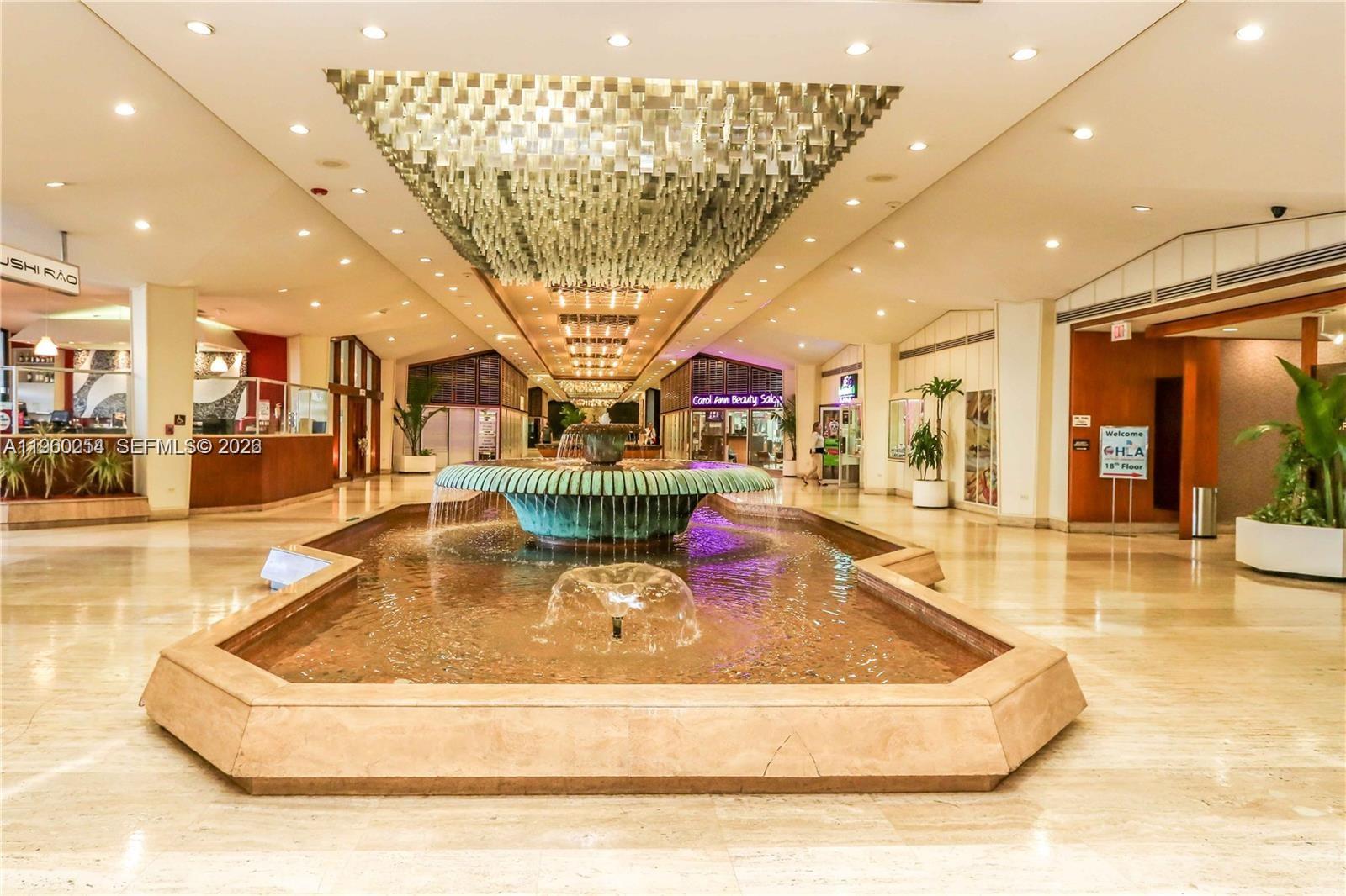 A large indoor lobby features a central tiered fountain beneath an expansive, decorative light fixture — 999 Brickell Bay Drive # 1202, Miami FL 33131, MIAMI, FL