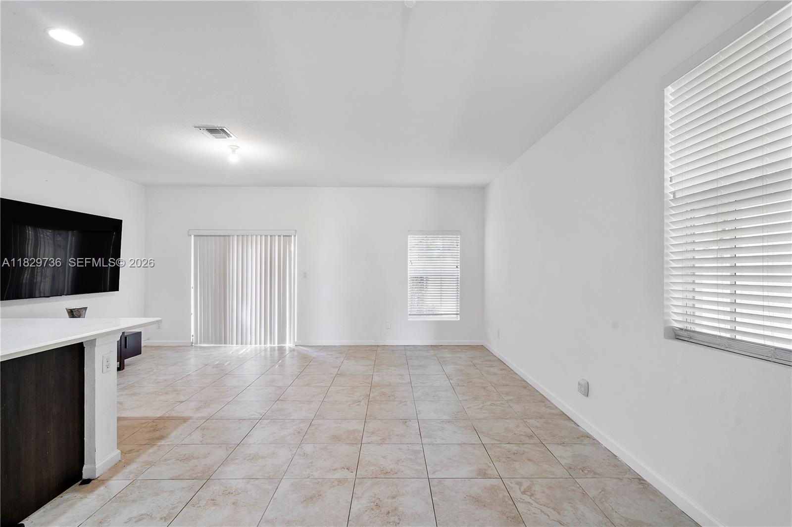 A bright, empty living room features white walls, beige tile flooring, and vertical blinds — 1128 NW 40th Ter, Lauderhill FL 33313, LAUDERHILL, FL