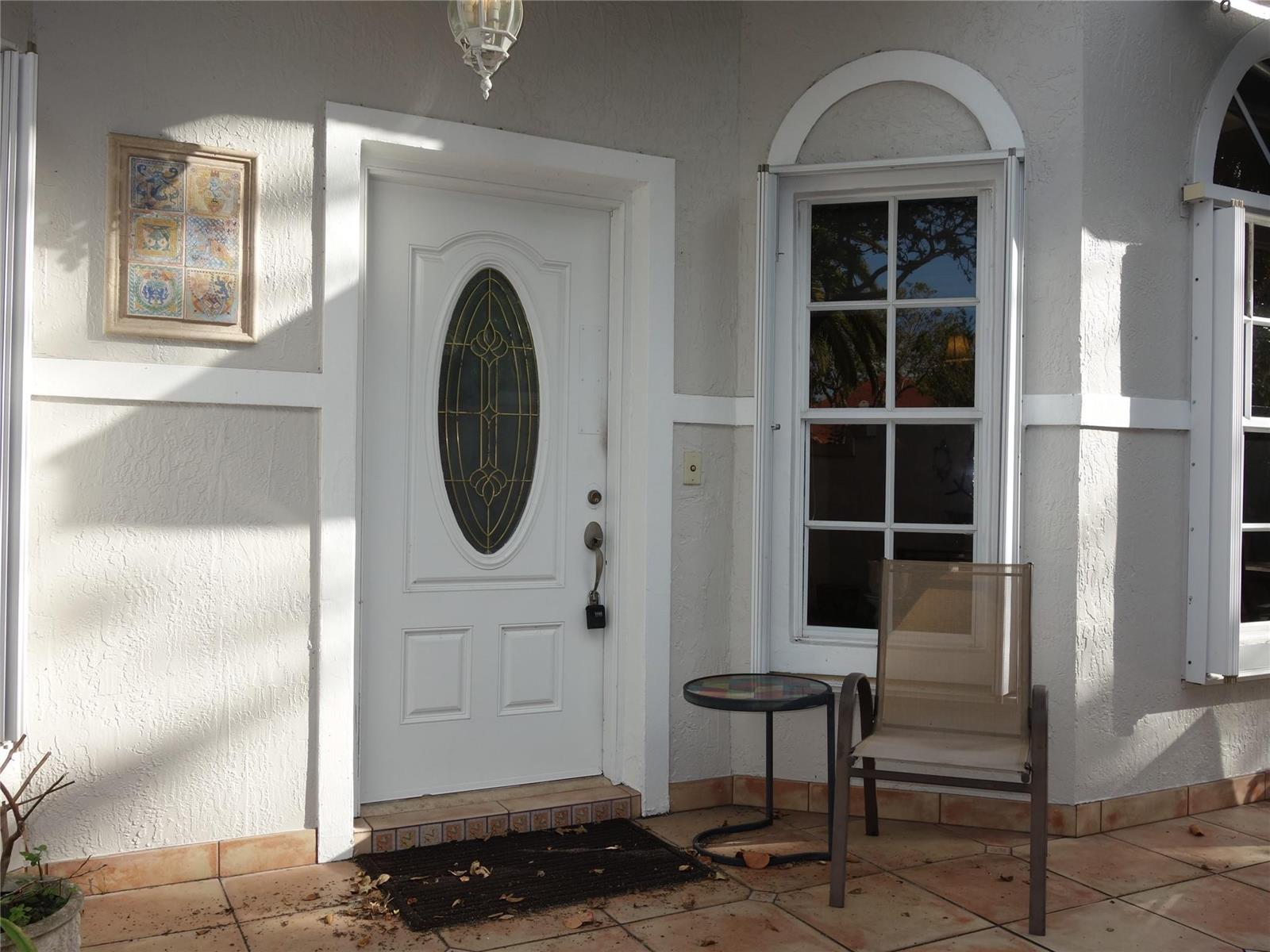 A white front door with an oval glass insert sits beside a window and chair — 4106 Carriage Drive # 5C, Pompano Beach FL 33069, POMPANO BEACH, FL