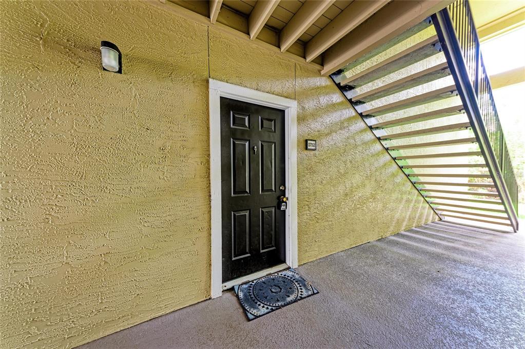 A black front door with a doormat sits under an outdoor staircase on a concrete patio — 4040 CROCKERS LAKE BOULEVARD UNIT 12, SARASOTA, FL
