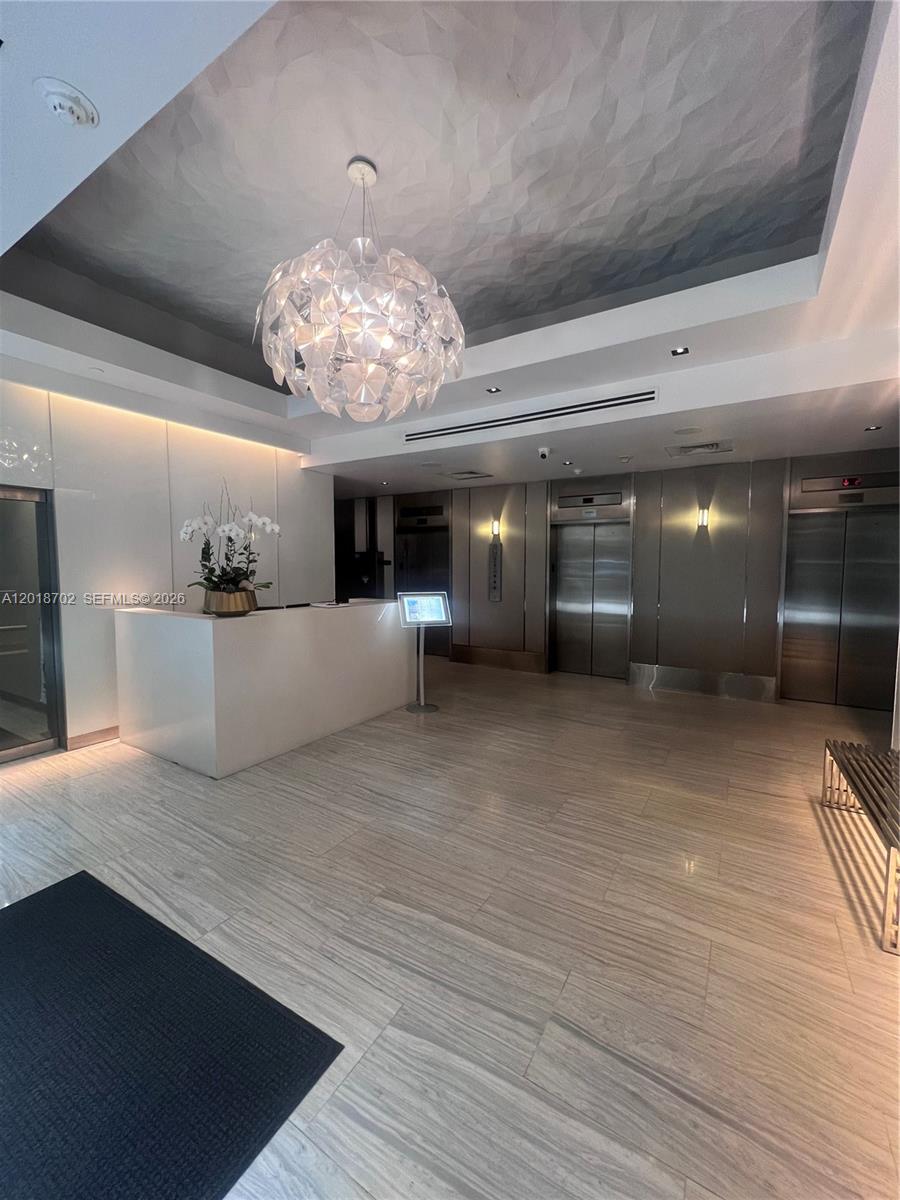 A modern building lobby features a white reception desk, metallic elevators, and a decorative chandelier — 150 SE 2nd Ave # 208, Miami FL 33131, MIAMI, FL