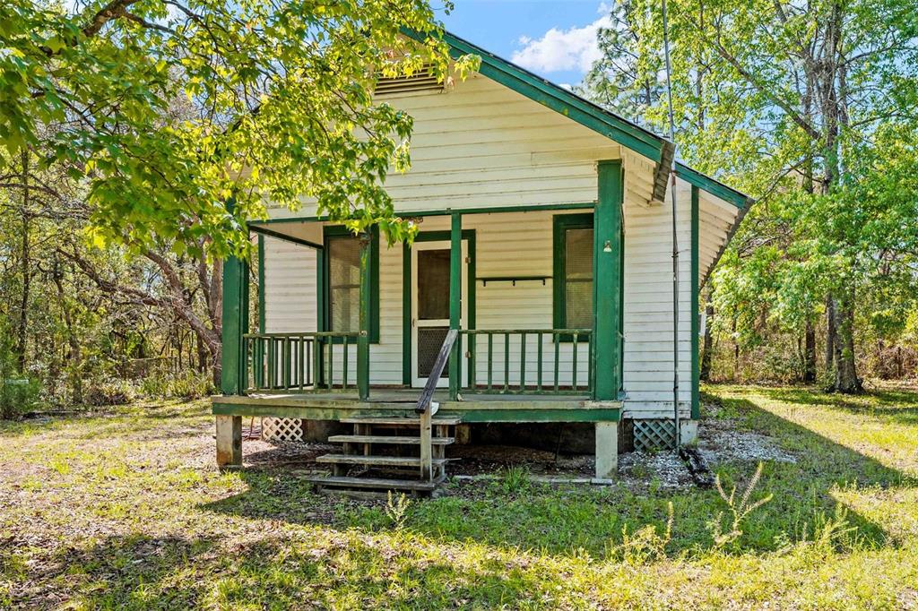 A small, weathered white cottage with a green-trimmed front porch sits in a wooded yard — 11317 SALINA STREET, BROOKSVILLE, FL