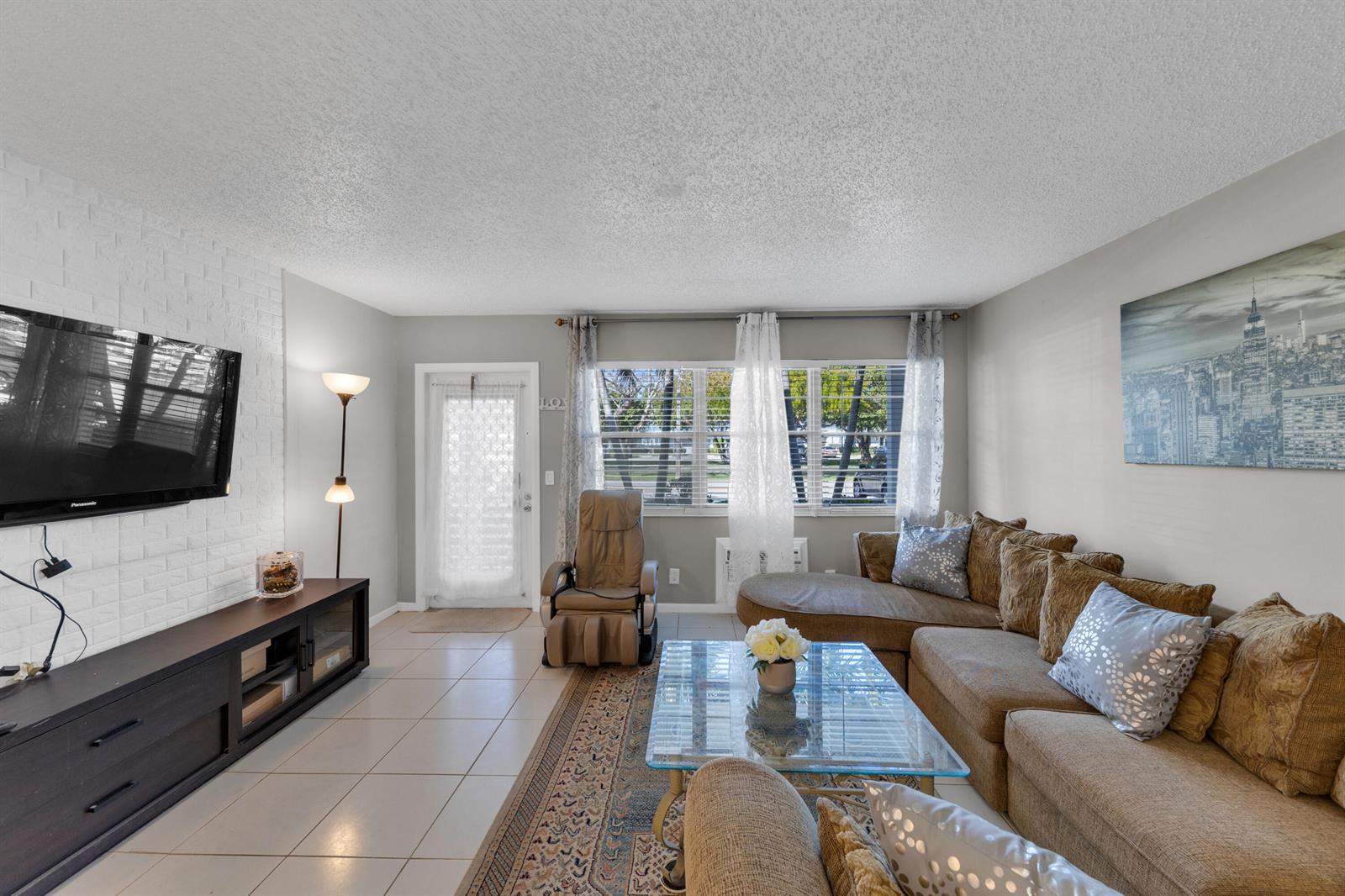 A living room features a brick accent wall, tile flooring, a sectional sofa, and windows — 5 Newport A, Deerfield Beach FL 33442, DEERFIELD BEACH, FL