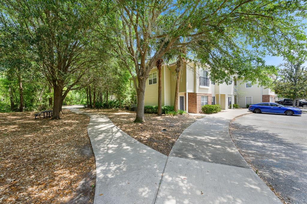 A concrete walkway leads to a multi-story apartment building surrounded by trees and a parking lot — 5062 NW 44TH LANE UNIT 102, GAINESVILLE, FL