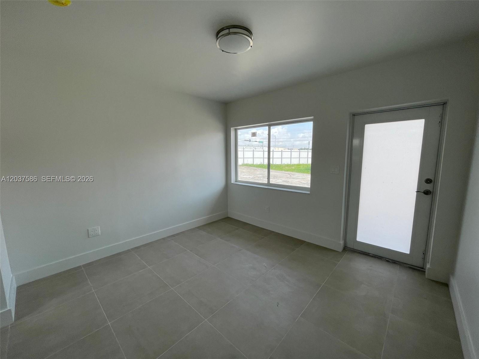 An empty room features white walls, large light-colored floor tiles, a window, and a door — 2196 Ali Baba Ave # 6, Opa-Locka FL 33054, OPA-LOCKA, FL
