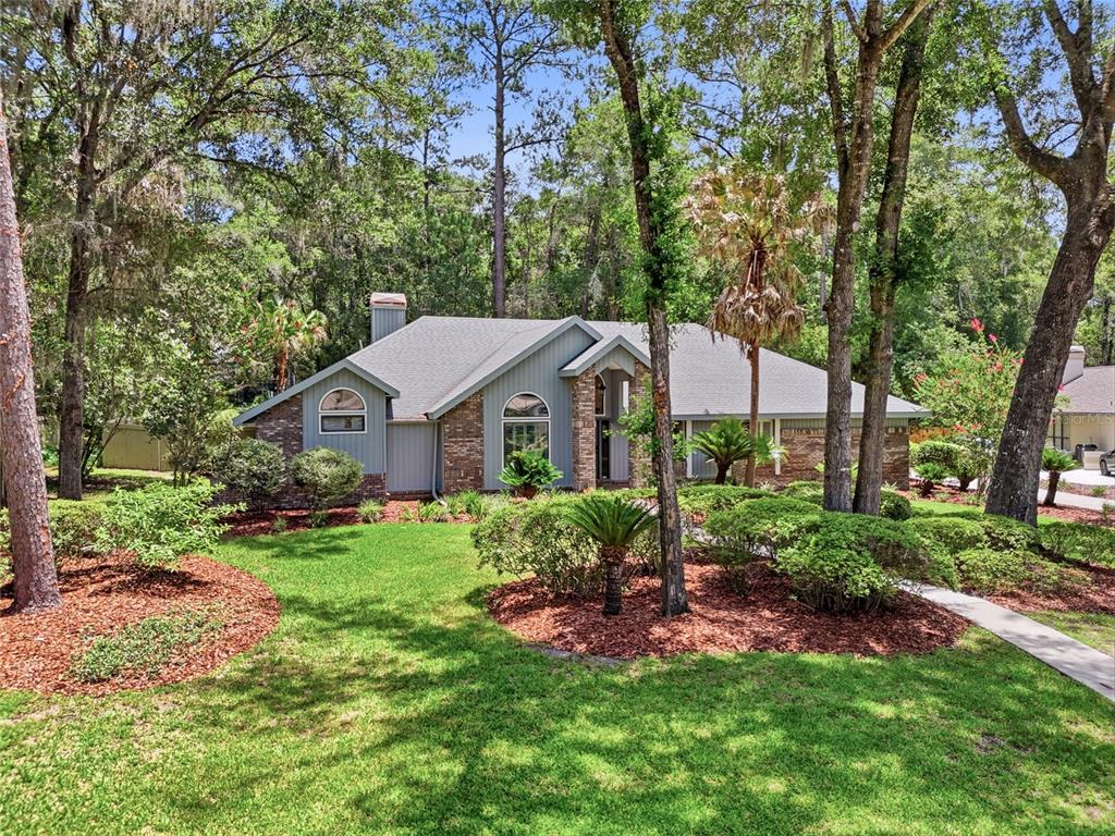A single-story house with brick and gray siding sits among trees and landscaped bushes — 8959 SW 44TH LANE, GAINESVILLE, FL