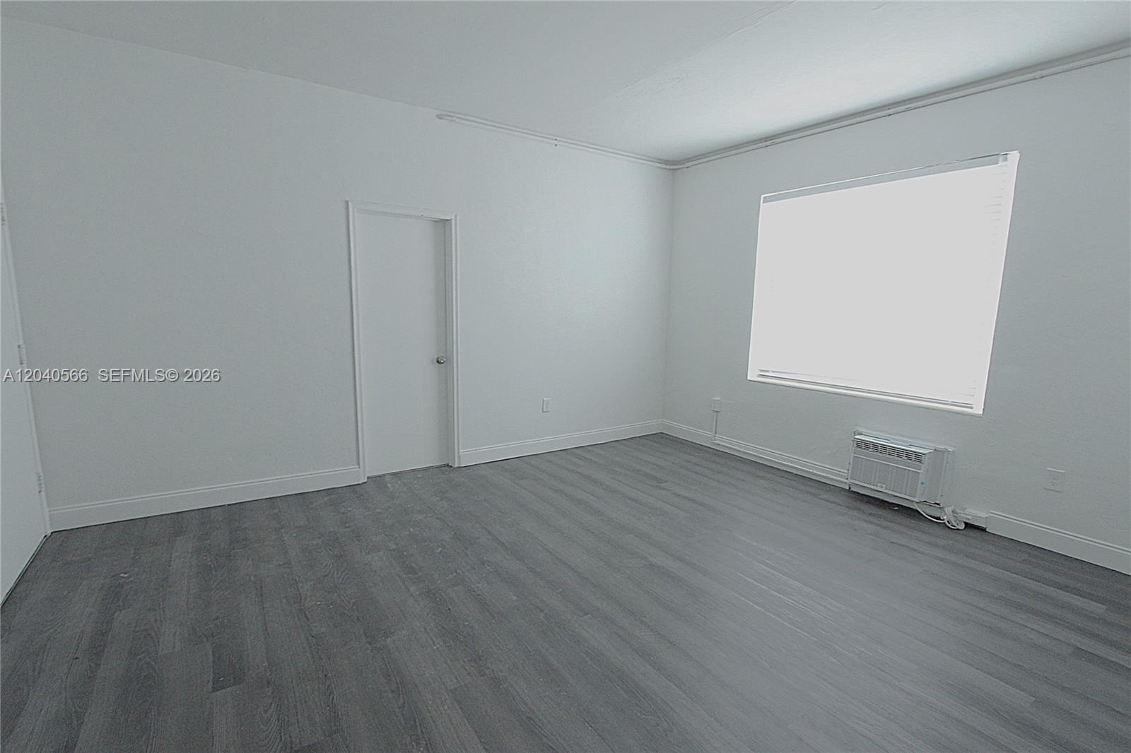 An empty room features white walls, gray wood-look flooring, a window, and a wall-mounted air conditioner — 1115 Euclid Ave # 11, Miami Beach FL 33139, MIAMI BEACH, FL