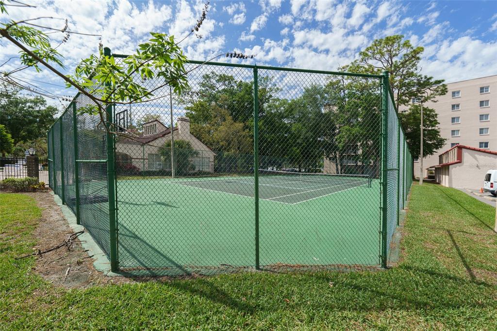 A fenced-in outdoor tennis court sits on a grassy lawn next to a building — 1150 CARMEL CIRCLE UNIT 302, CASSELBERRY, FL