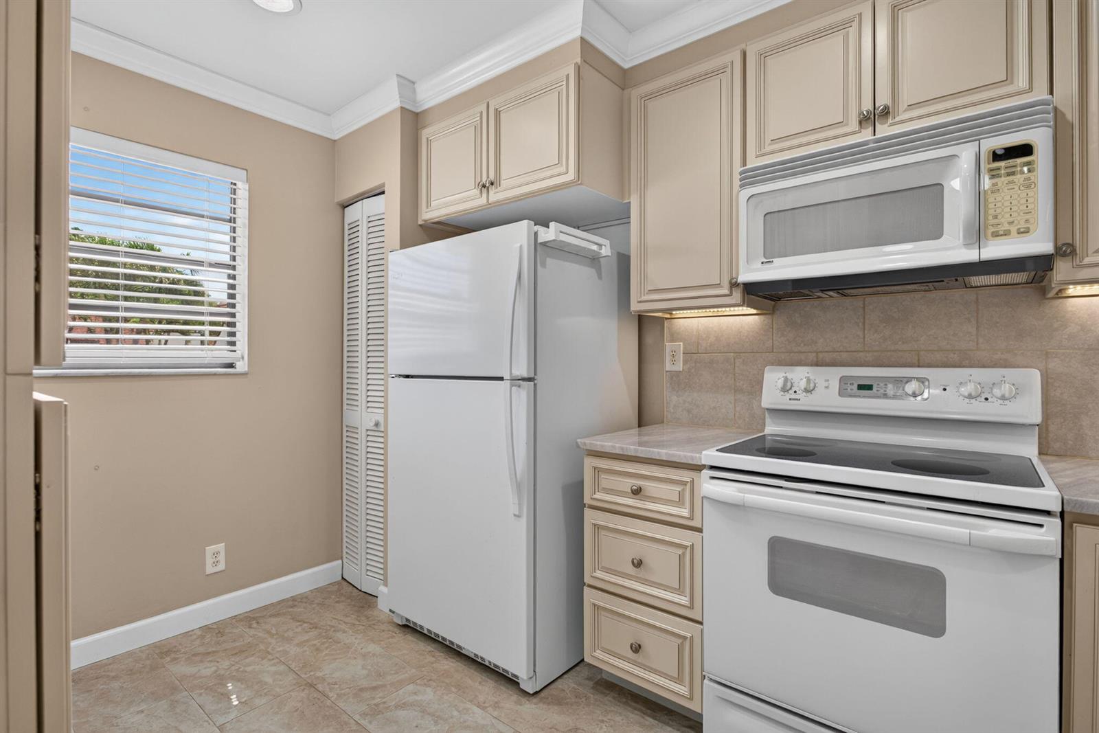 A kitchen features beige cabinets, white appliances, a tiled backsplash, and a window with blinds — 353 NW 36th Avenue, Deerfield Beach FL 33442, DEERFIELD BEACH, FL