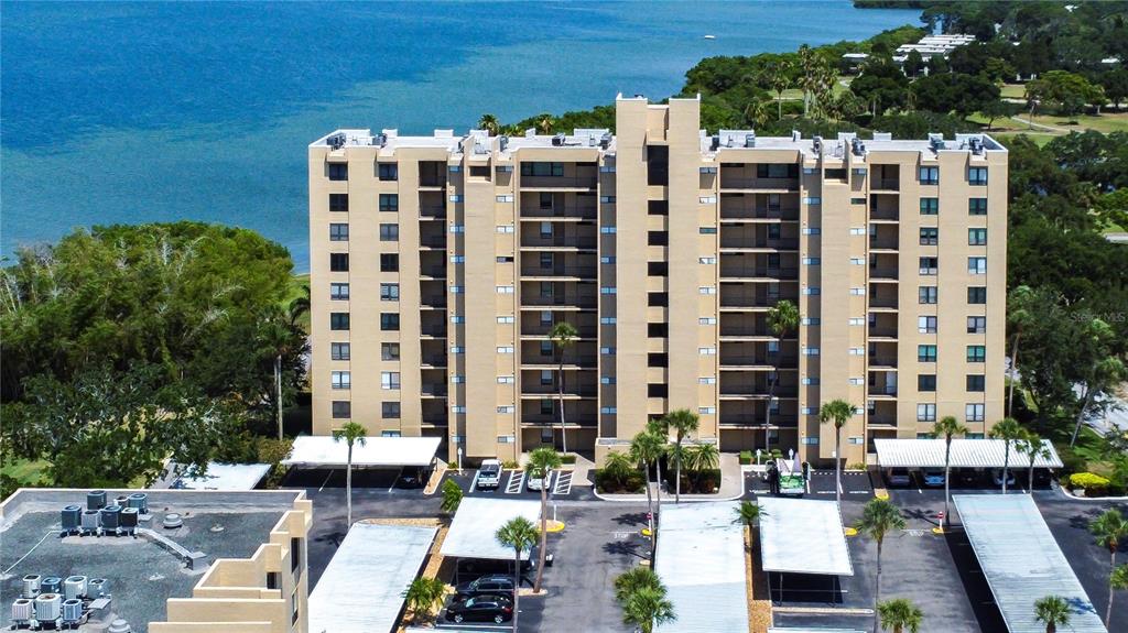 An aerial view shows a tall, tan apartment building with covered parking spaces and water — 2616 COVE CAY DRIVE UNIT 506, CLEARWATER, FL