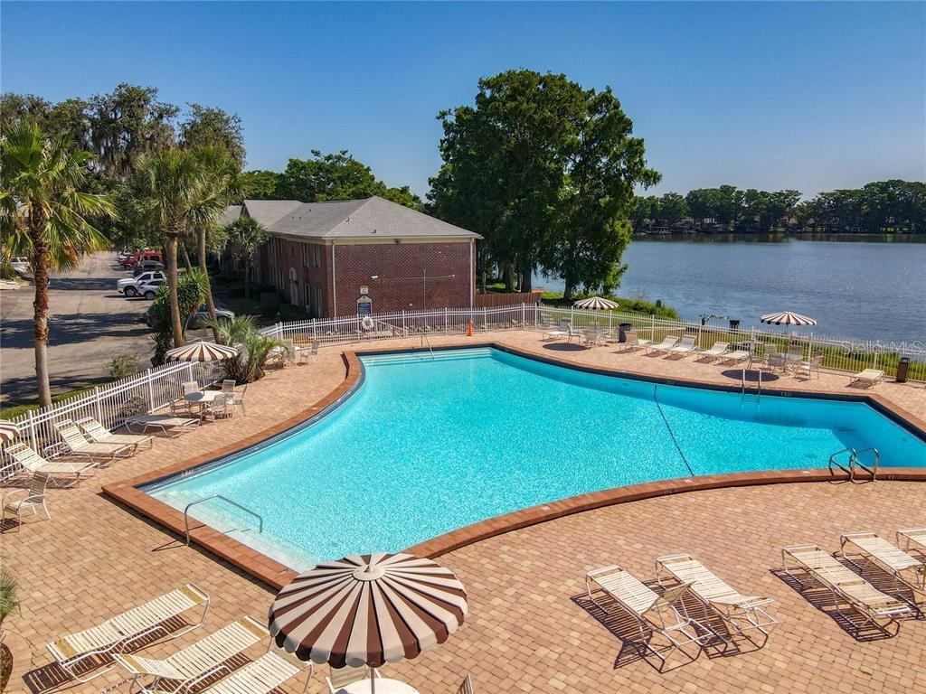An outdoor swimming pool with lounge chairs and umbrellas overlooks a lake and building — 116 GEORGETOWN DRIVE, CASSELBERRY, FL