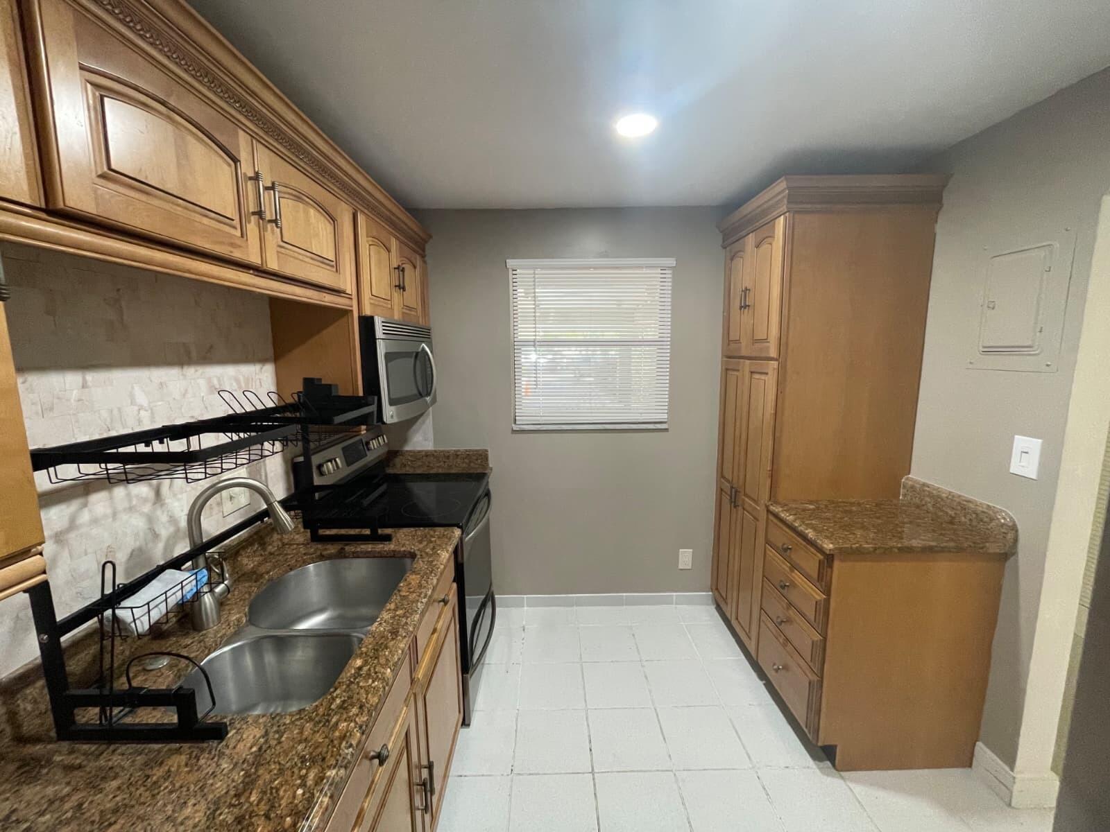A kitchen features wood cabinets, granite countertops, a double sink, and white tile flooring — 2322 S Cypress Bend Drive # 110, Pompano Beach FL 33069, POMPANO BEACH, FL