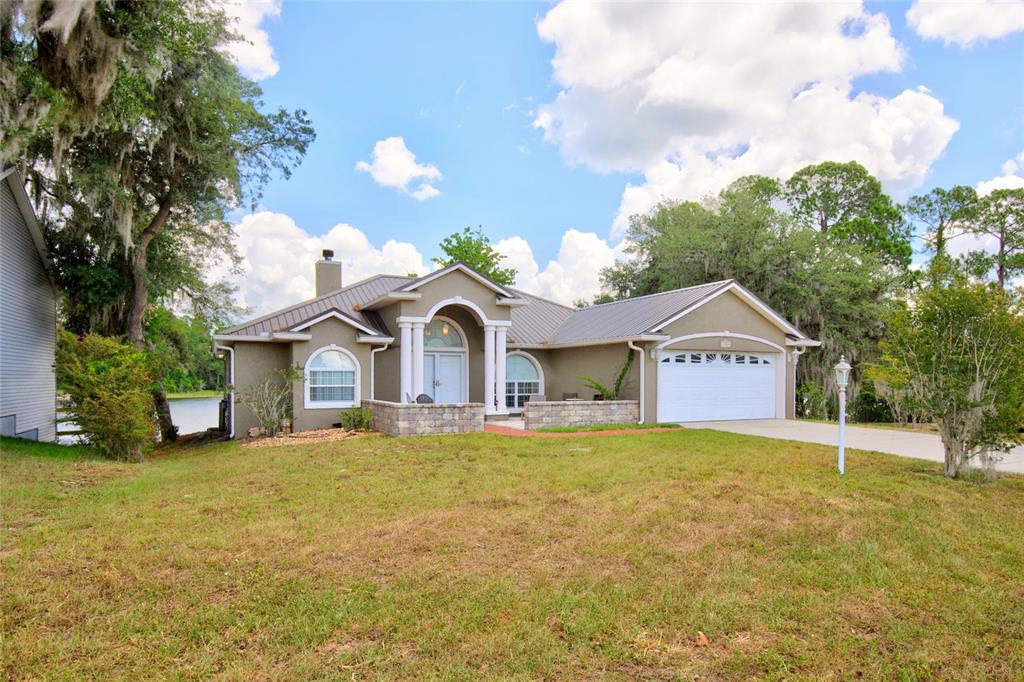 A tan single-story house with a white garage door and arched entryway sits behind a driveway — 13059 NE 7TH LOOP, SILVER SPRINGS, FL