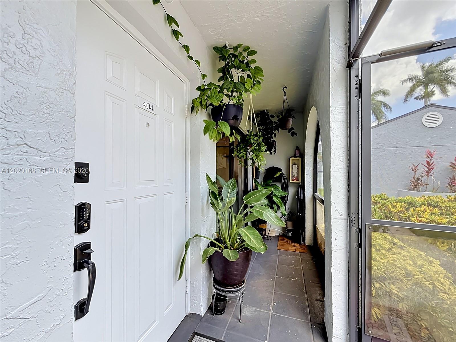 A white front door with a digital lock opens to a tiled entryway with potted plants — 18296 Mediterranean Blvd # 404, Hialeah FL 33015, HIALEAH, FL